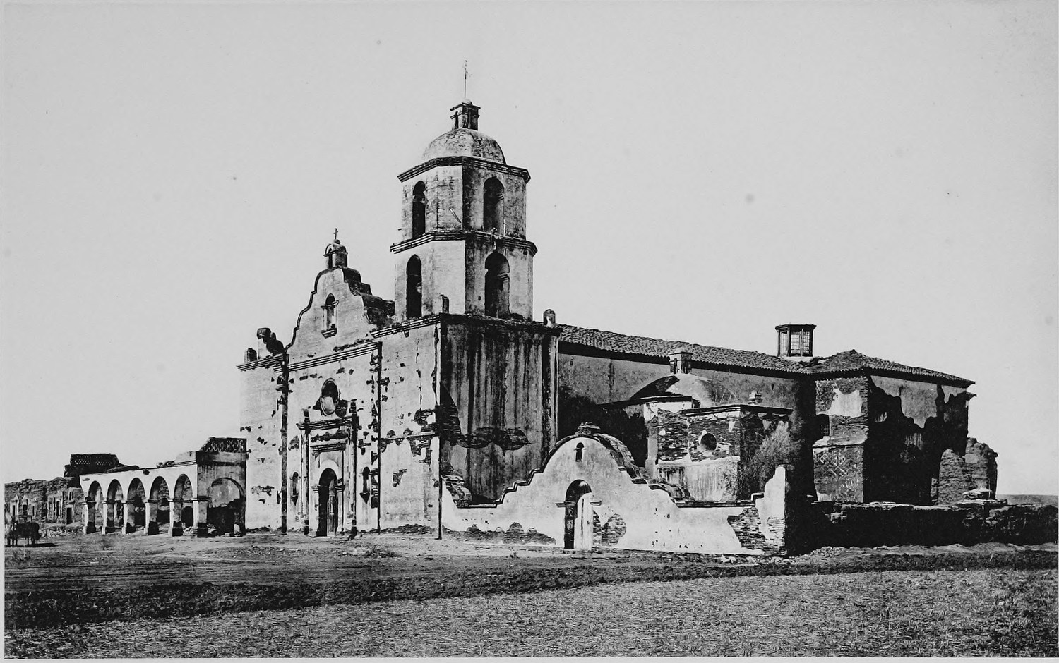 A mission church with a domed bell tower, a long arcade of arches running off to the left and sections of wall in ruins