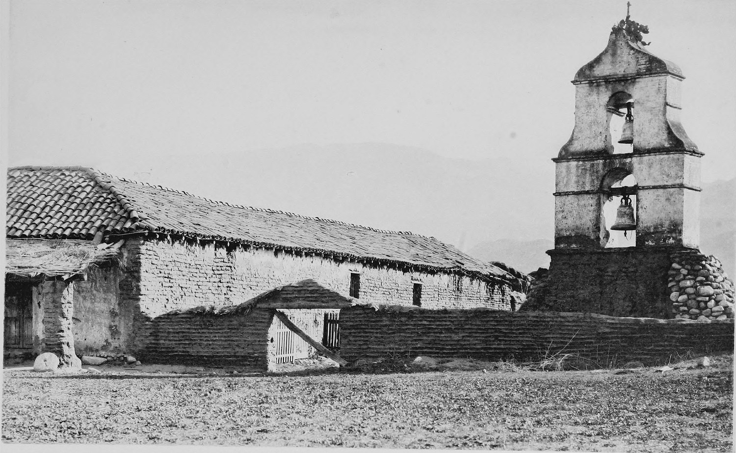 A freestanding stone bell tower holding two bells, beside a long tile roofed adobe building on dry ground