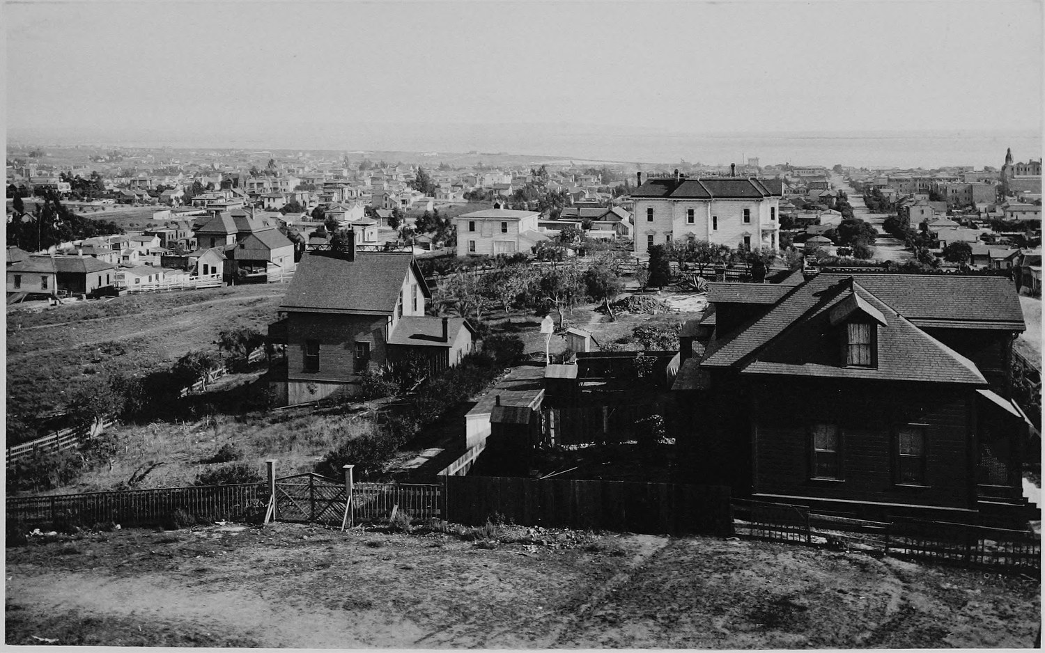 A town of frame houses seen from a hillside, a dark shingled house in the foreground, open water on the horizon