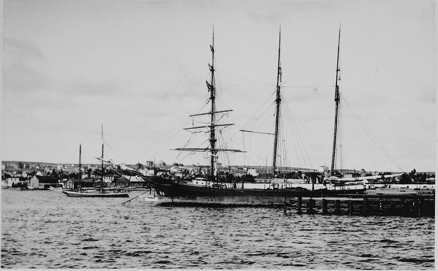 A three masted sailing ship moored at a wharf with smaller schooners alongside, the low town along the shore behind and choppy water in front