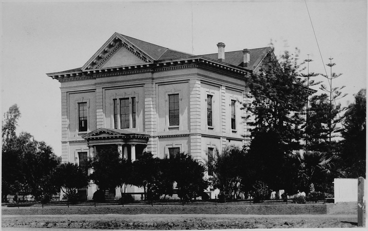 A two story courthouse with a pedimented portico and an iron fence, young conifers at one side and a dirt street in front
