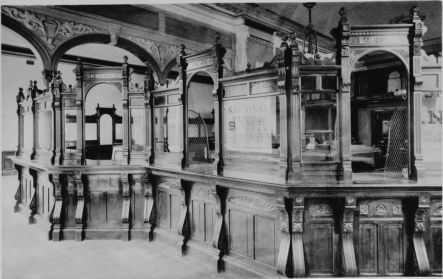 The inside of a bank, a long carved wooden counter with grilled teller windows and ornate arches overhead