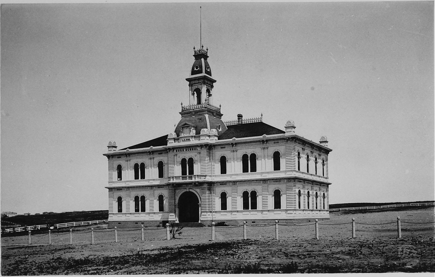 A large three story school building with a cupola and arched windows, standing alone behind a post and rail fence on bare ground