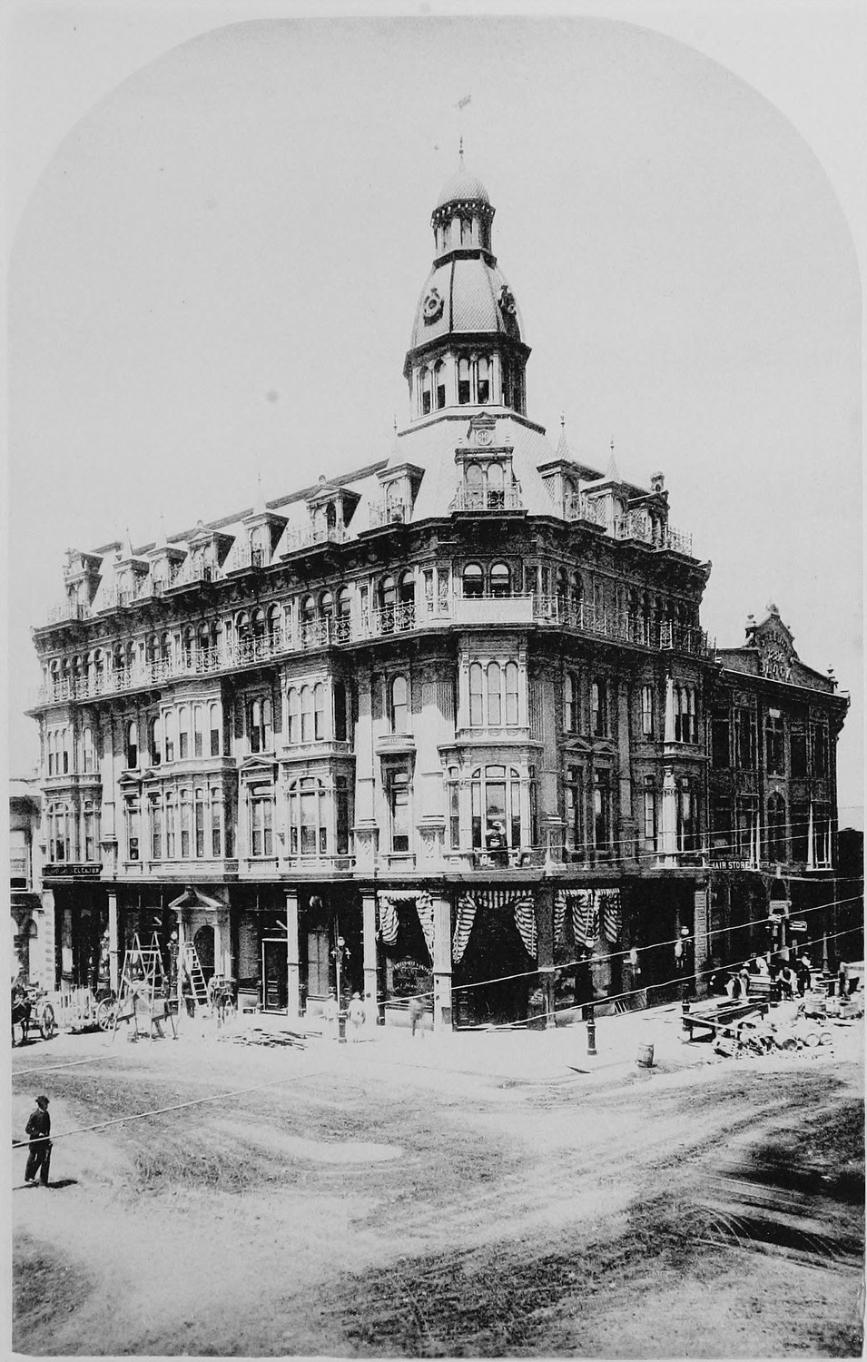 A four story commercial block on a street corner, bay windows up every floor and a domed corner tower, shops at street level