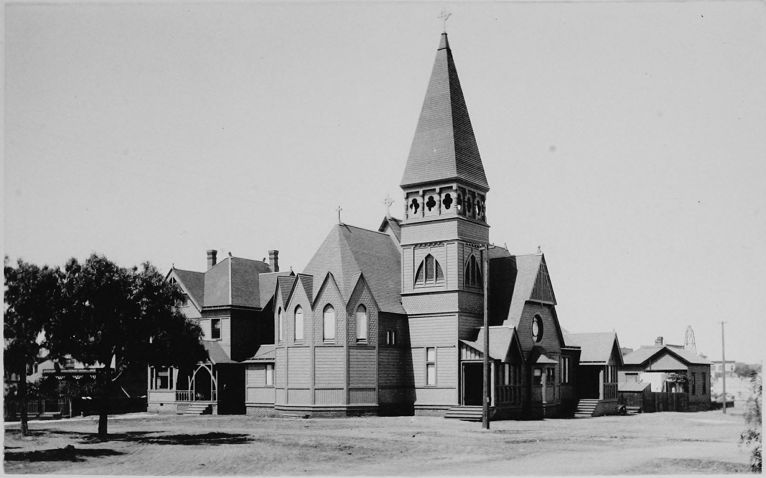 A wooden Gothic Revival church with a tall shingled spire, smaller gabled buildings beside it and a wide dirt street