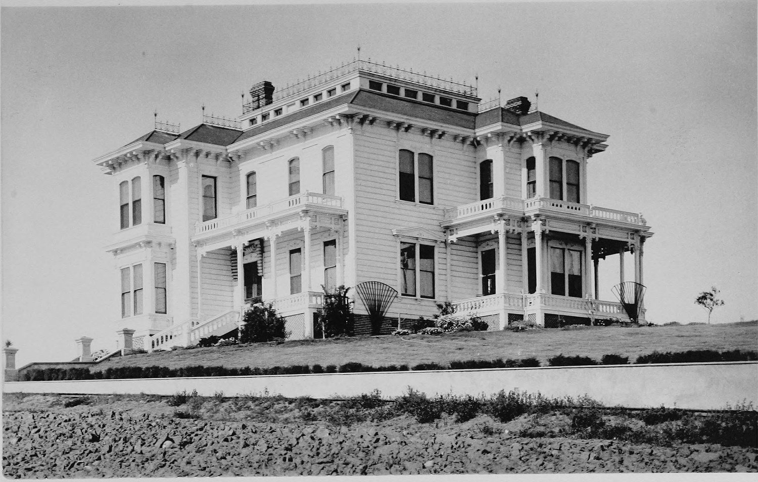 A large house with a balustraded flat roof, bay windows and a verandah, standing on a bare lot above a dirt road