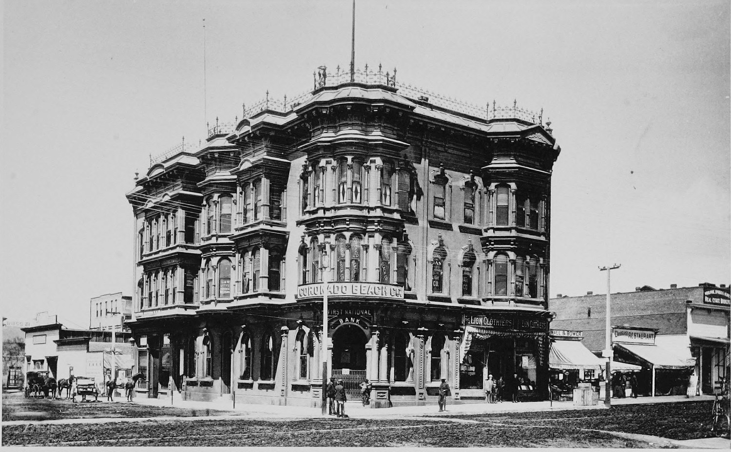 A four story corner bank building with tiers of bay windows, awnings over the ground floor shops, horses and wagons in the dirt street