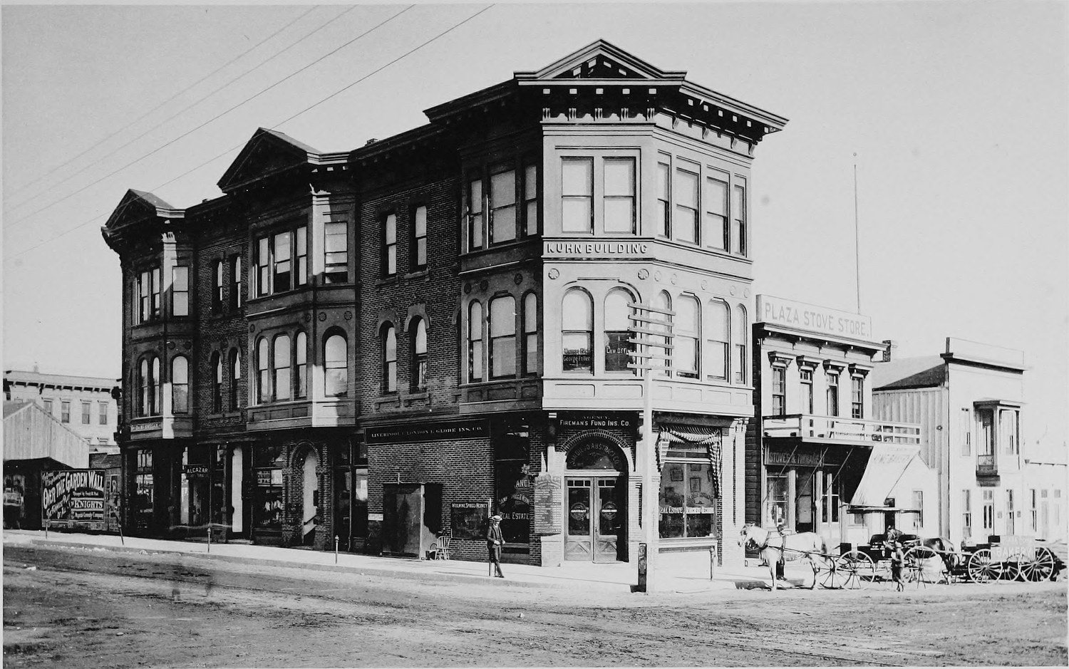 A three story brick commercial building on a corner, a real estate office at street level, people and buggies along the sidewalk