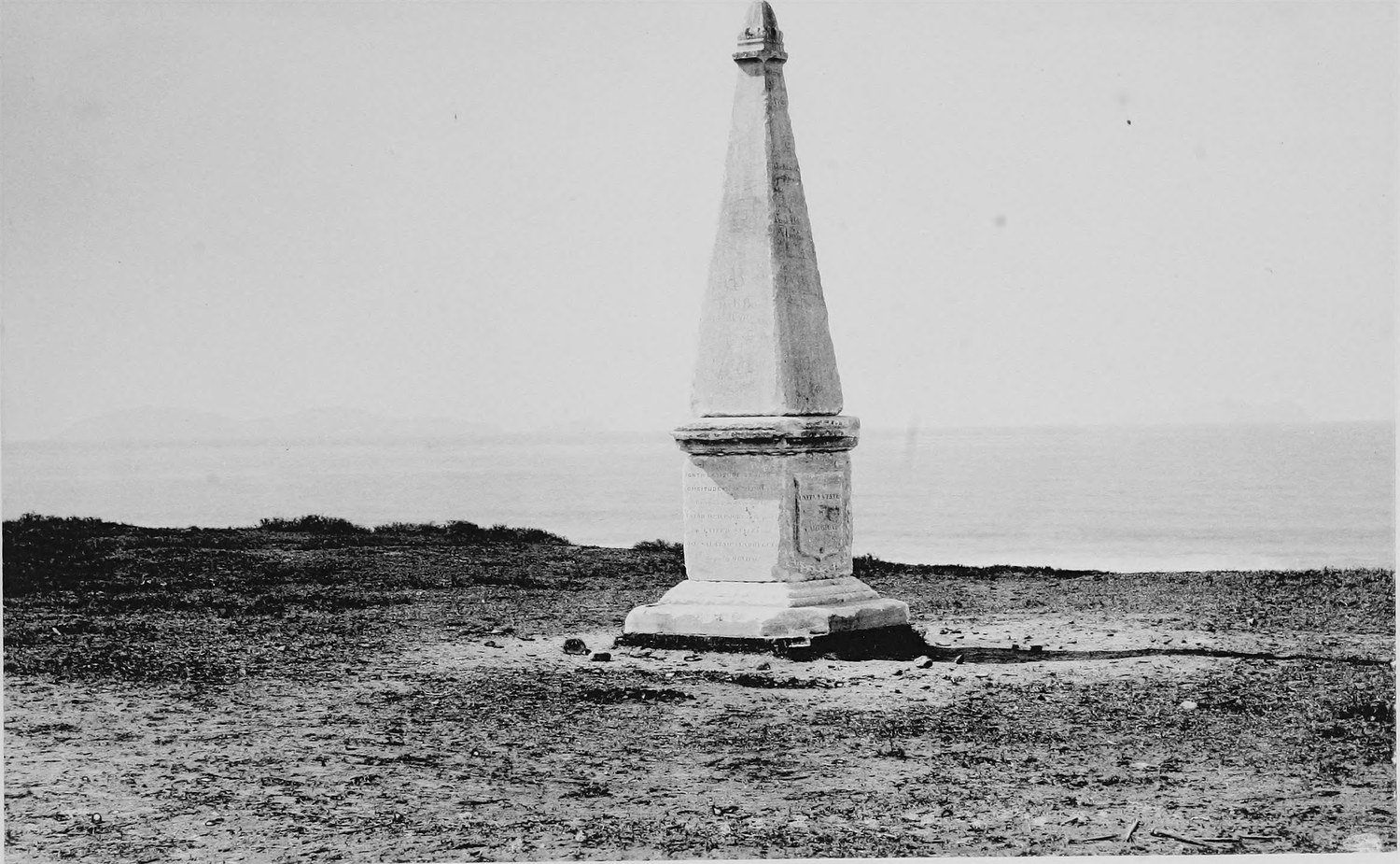 A tall white stone obelisk standing alone on a low bluff above the sea