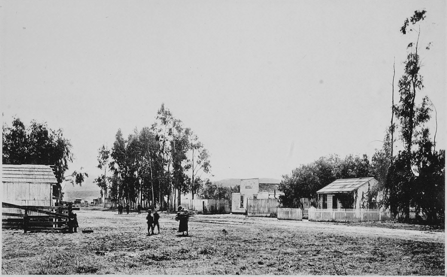 A dirt crossroads with a few board buildings and a fenced yard, tall thin trees along the road and several people standing in the open