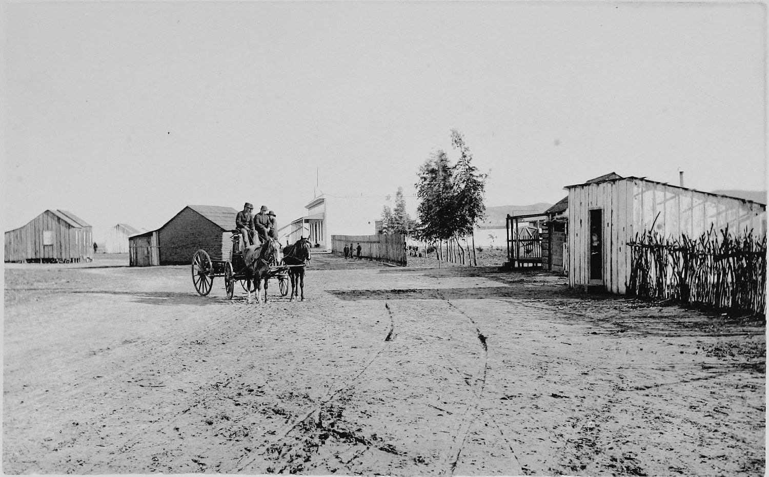 A wide sandy street with a horse and buggy, low board and adobe buildings along both sides and a cane fence at the right