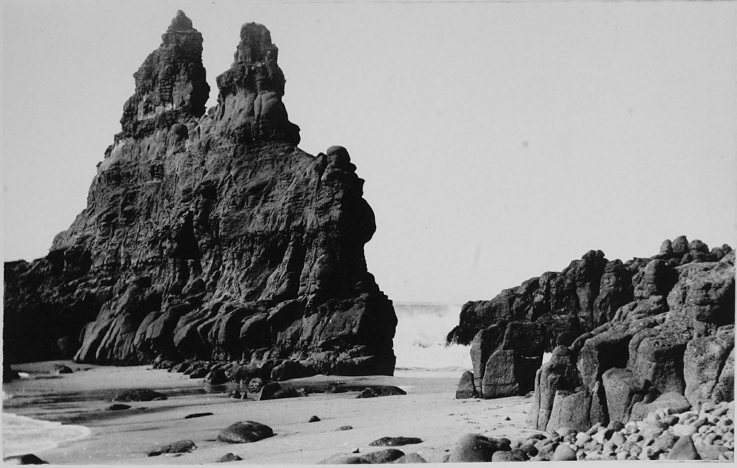 A tall jagged sea stack rising off a sand and cobble beach, a second lower rock beside it