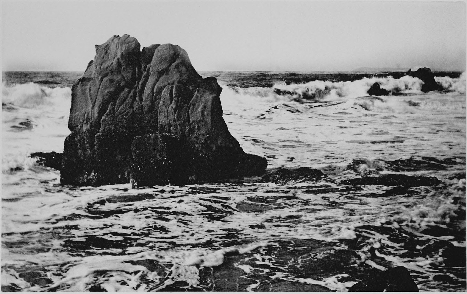 A single blocky sea stack standing in breaking surf, more rock showing through the water in front