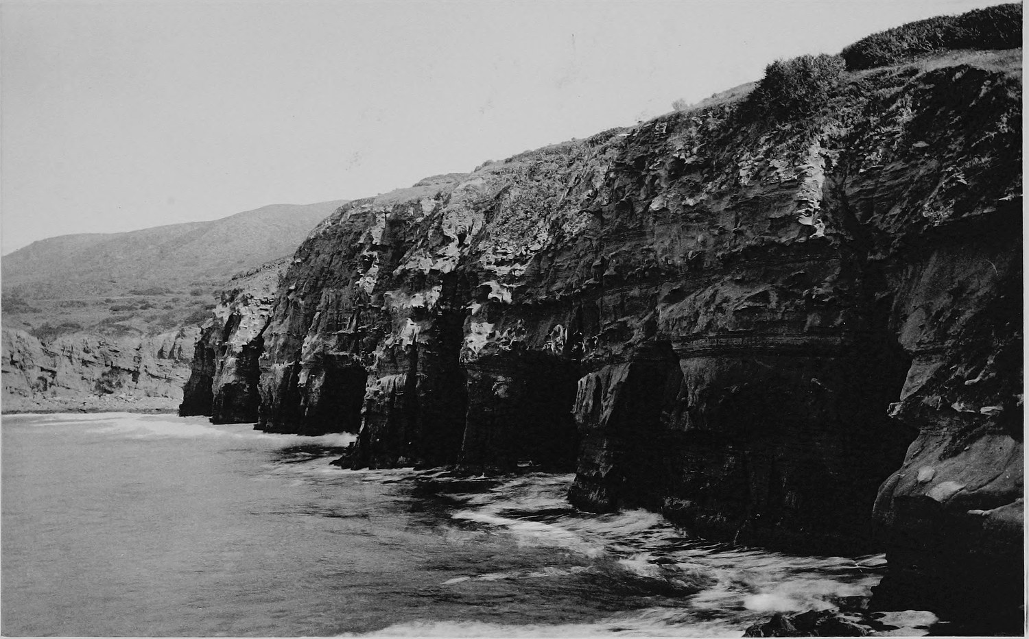 High layered sea cliffs curving away above a narrow beach, surf running along their base