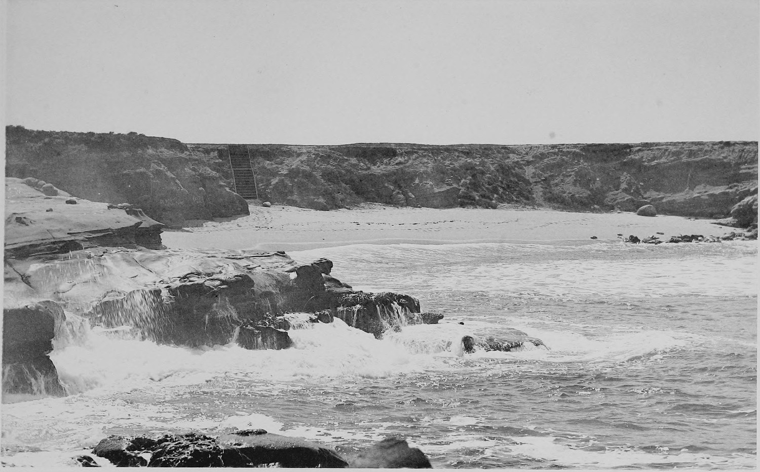 Surf breaking over a low rock shelf in the foreground with a long sandy beach and cliffs behind
