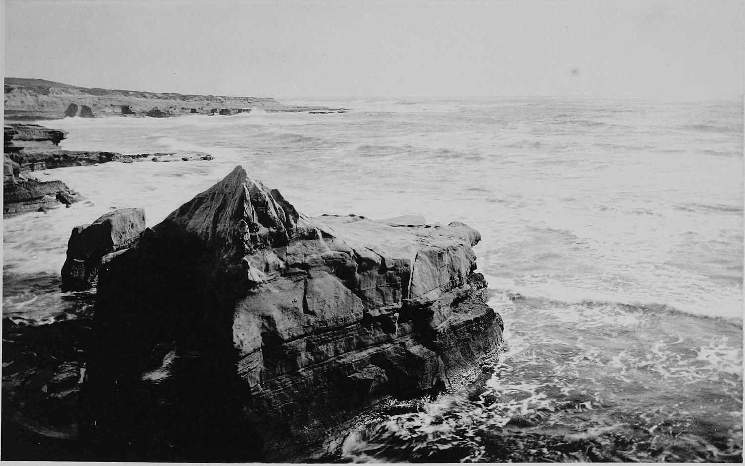 A large flat topped rock standing just off the shore with surf around it, low cliffs running away in the distance