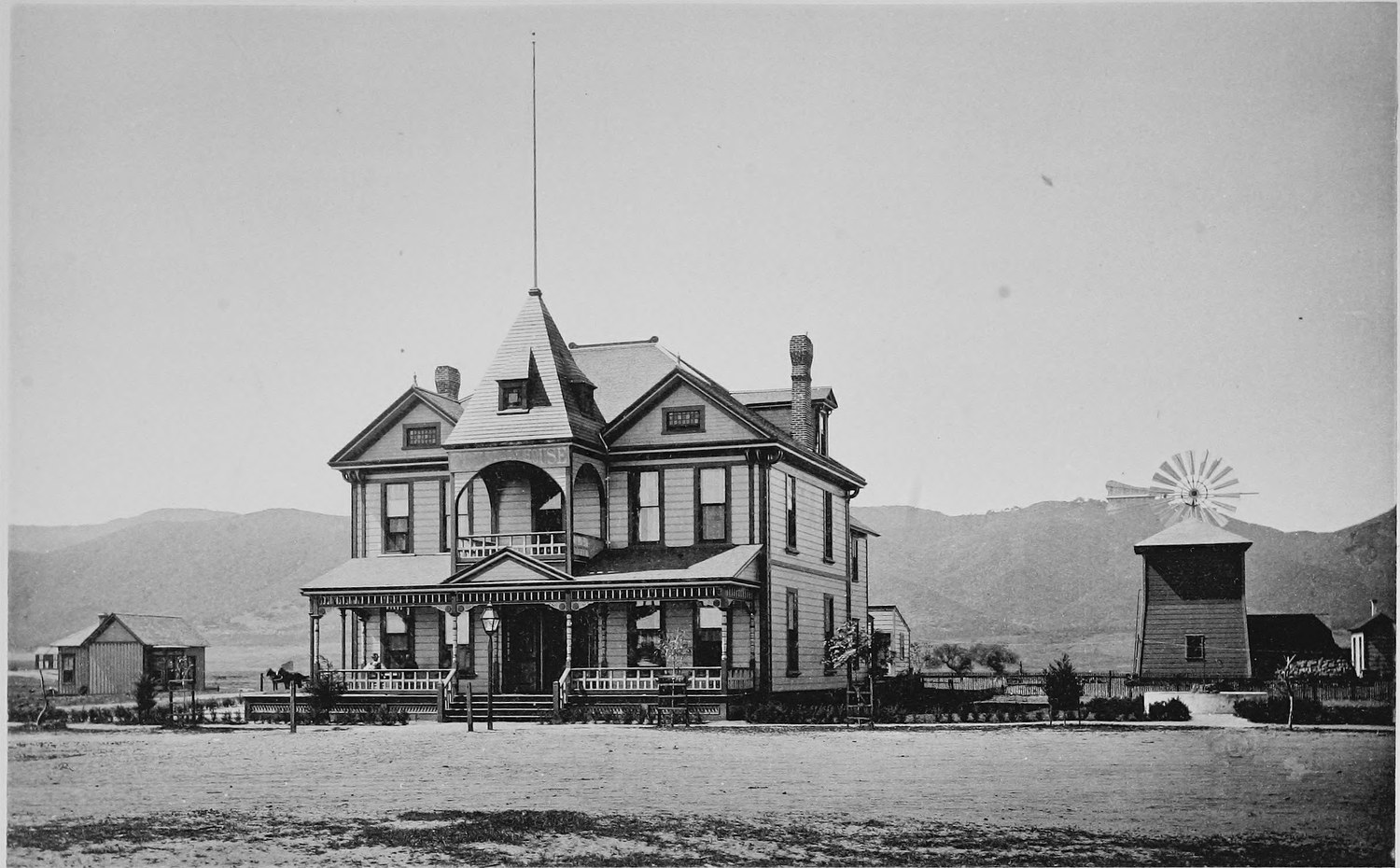 A large ornate wooden hotel with a tower and wide porches, a windmill and water tank at one side and mountains behind