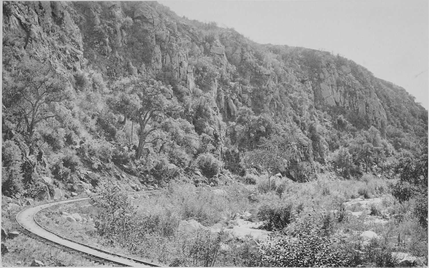 A single railway track curving along the floor of a steep brush covered canyon
