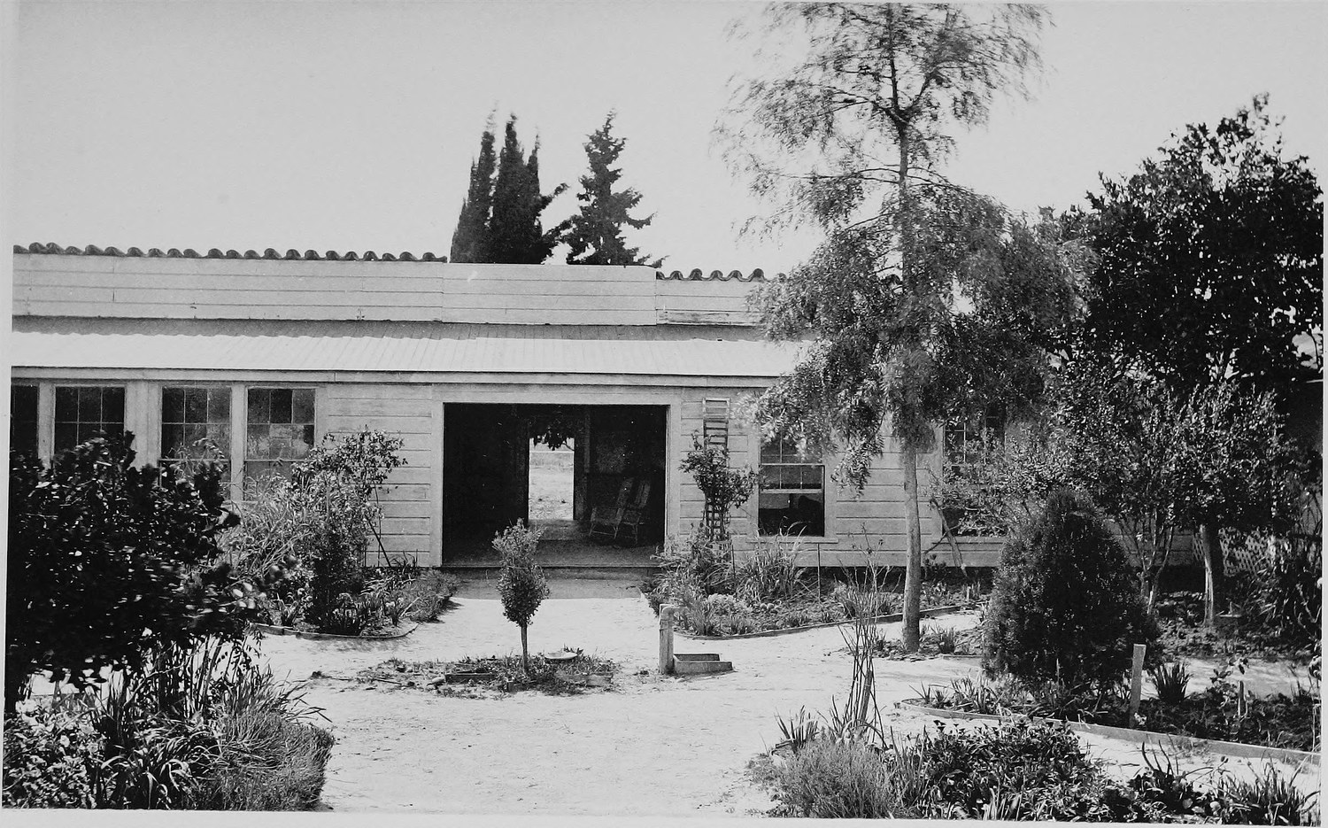 A single story house with a tiled roof seen across its garden, a gravel path to the open front door, cypress and pepper trees around it