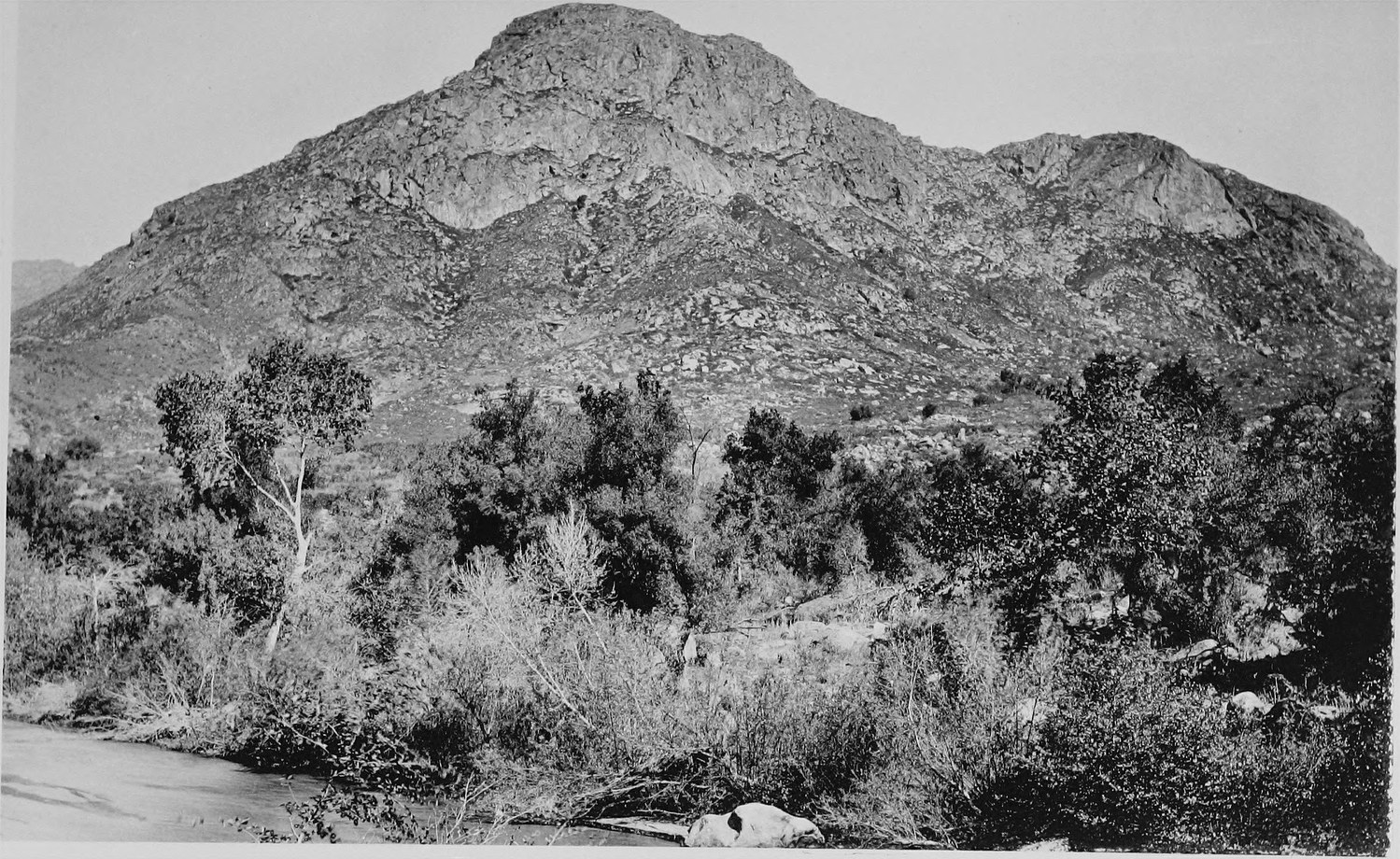 A steep rocky peak rising above brush and a sandy wash in the foreground