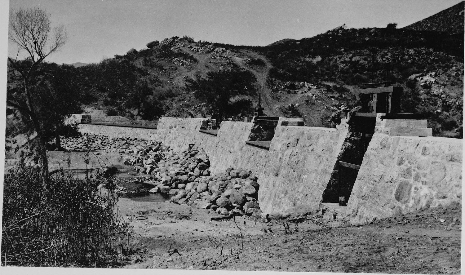 A low masonry diversion dam built across a rocky creek bed, loose stone piled against it and a hillside behind