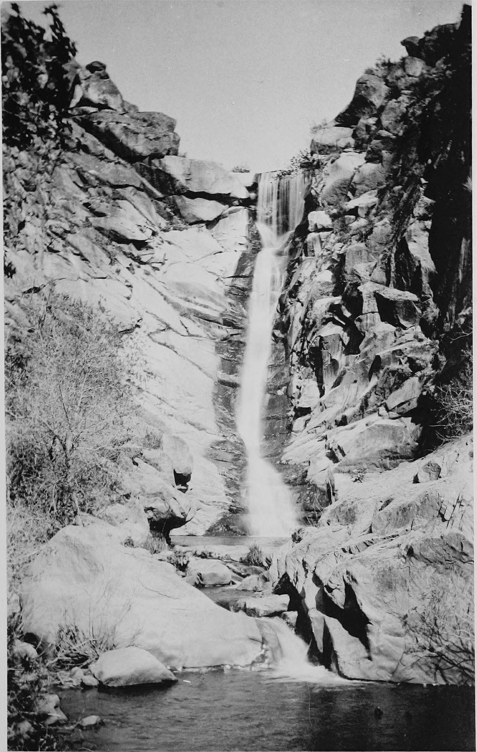 A waterfall dropping down a bare rock face into a narrow gorge, broken boulders and brush below