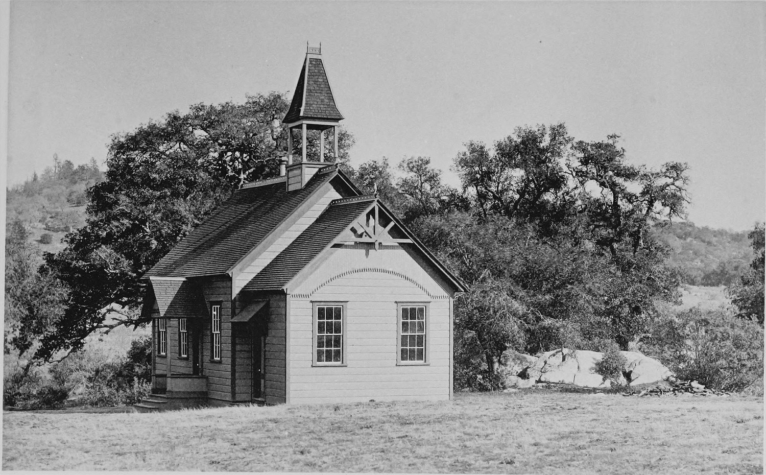 A white wooden schoolhouse with a bell tower and arched trim, oaks on both sides and open grass in front
