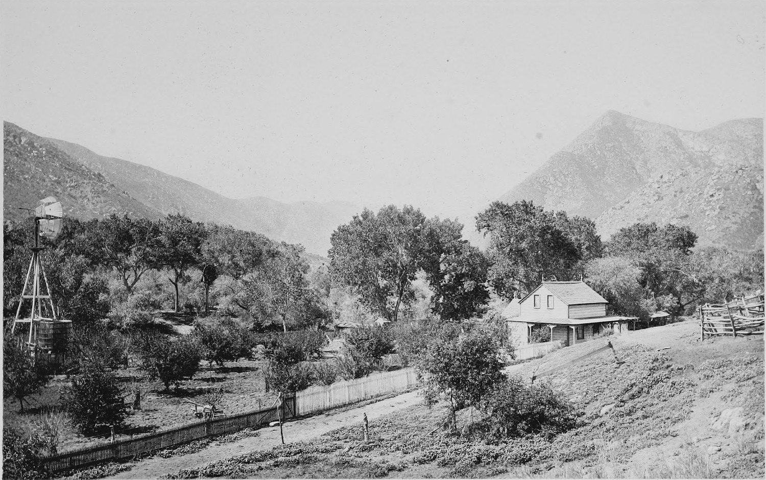 A valley with a windmill at the left, a farmhouse and fenced yard among trees, and a sharp peak behind