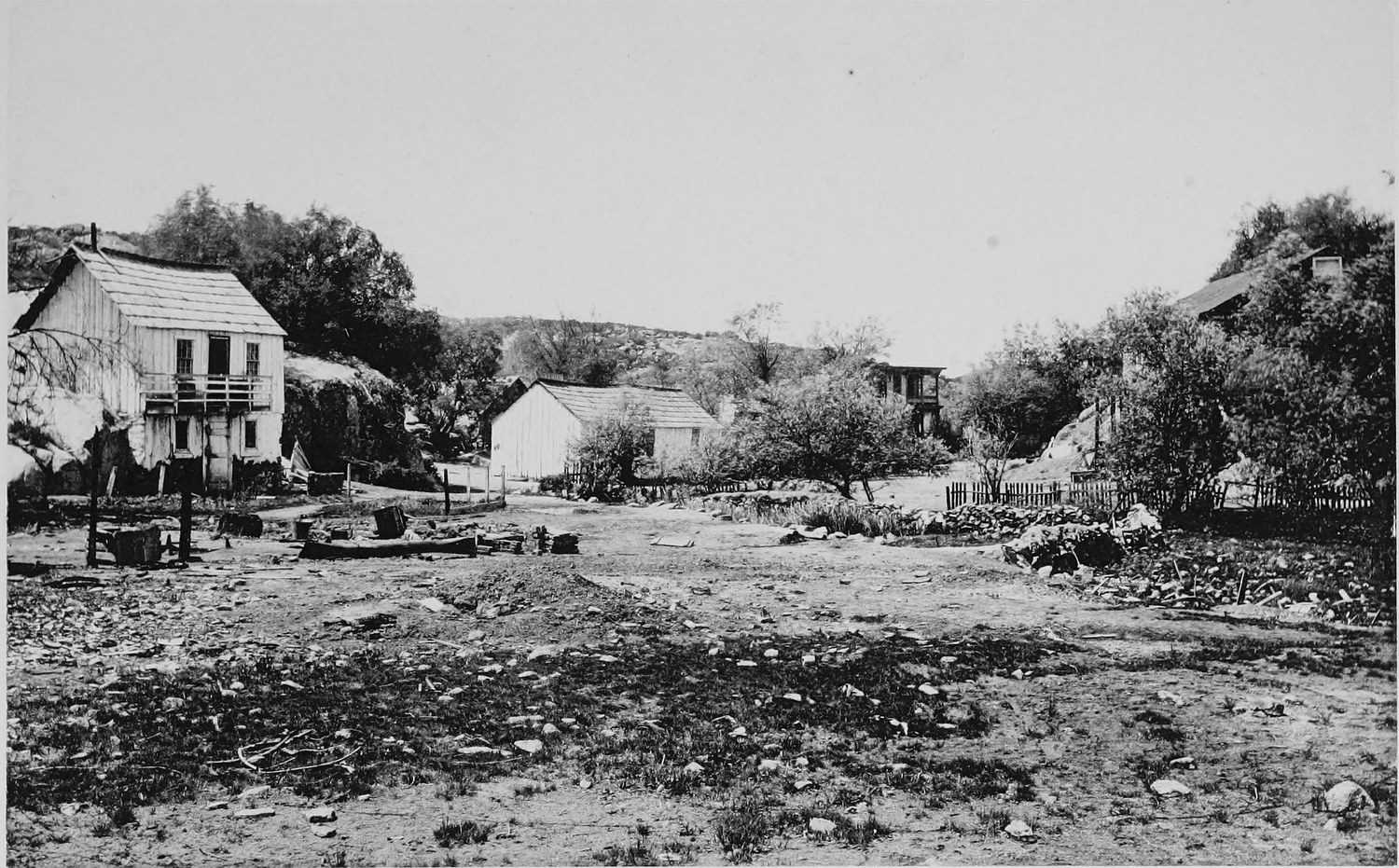A wide dirt street with a two story board hotel at the left, scattered houses and sheds, and brushy hills behind