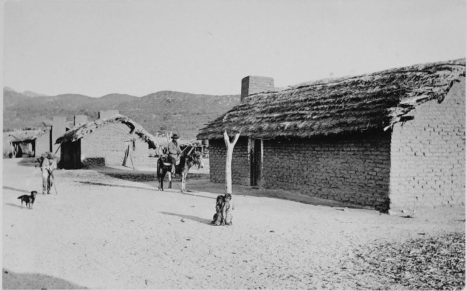 A low adobe building with a thatched roof on a bare dirt street, a man leading a horse past it and children standing nearby