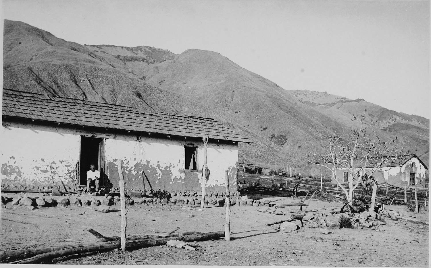 A long low adobe building with a tiled roof and a porch, a person sitting in the doorway, a bare rounded hill behind
