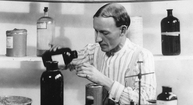 A thin man in a striped shirt pouring liquid from a dark bottle into a jar, with chemical bottles on a shelf behind him