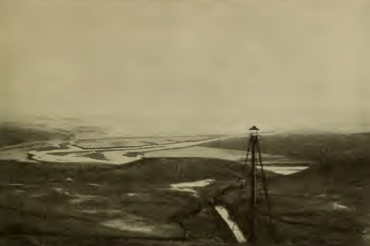 A drained valley floor with a winding stream and a tall steel gauge tower in the foreground