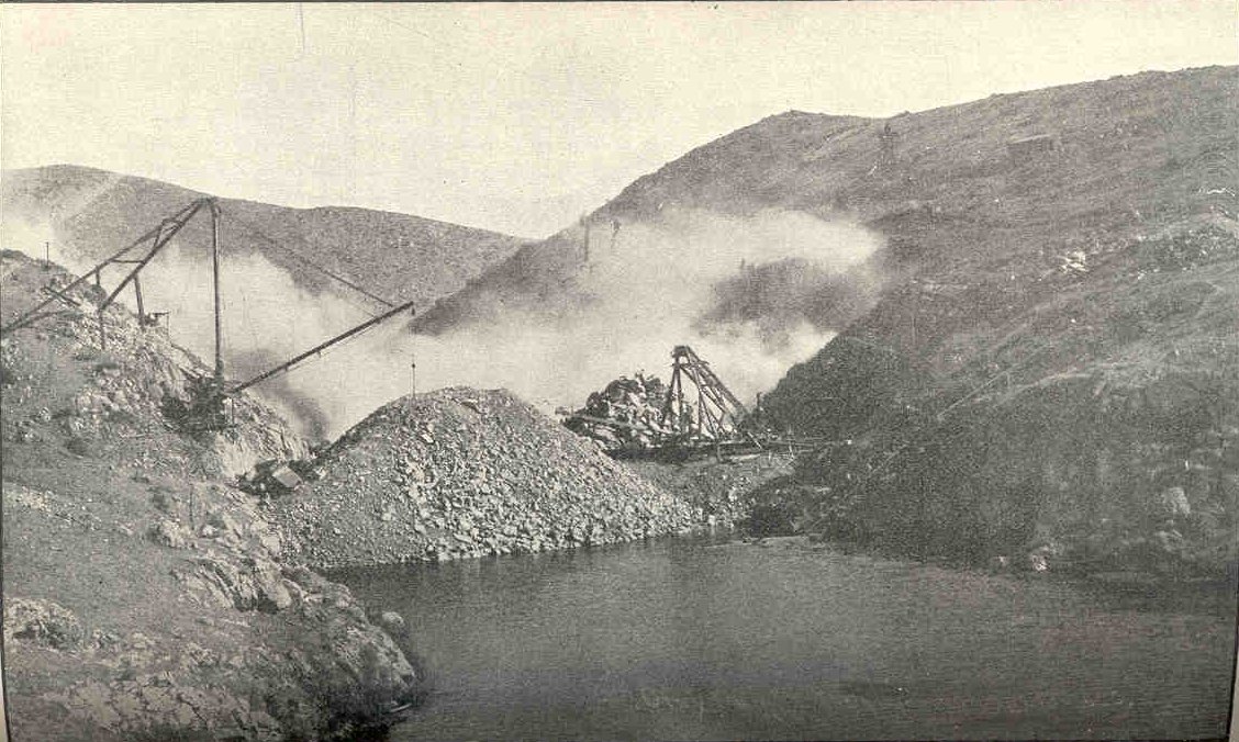 A cloud of dust rising from a canyon wall above a pool of water, with wooden derricks and a pile of broken rock