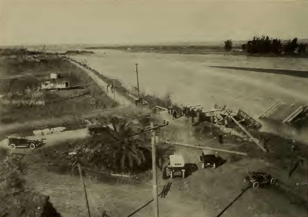 A wide flooded river with a broken wooden railroad trestle and road bridge, cars and a house on the near bank