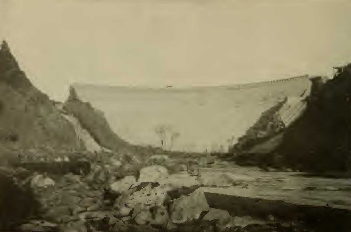 A curved masonry dam with rubble and boulders piled in the riverbed below it