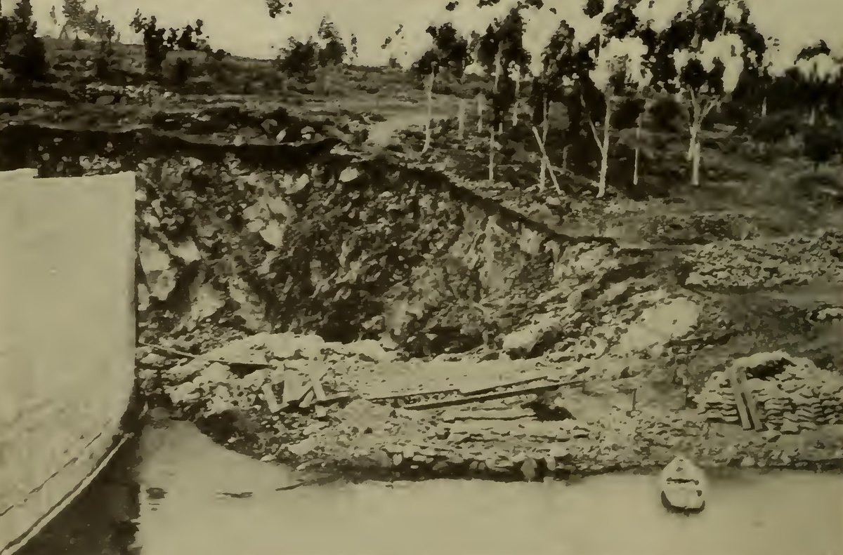 A deep washed out gap in the earth beside the end of a dam wall, with eucalyptus trees above