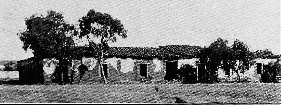 A long single story adobe house with a tile roof behind trees along a dirt street