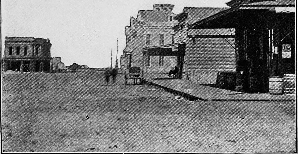 A wide dirt street with a few two story buildings and a store with barrels out front