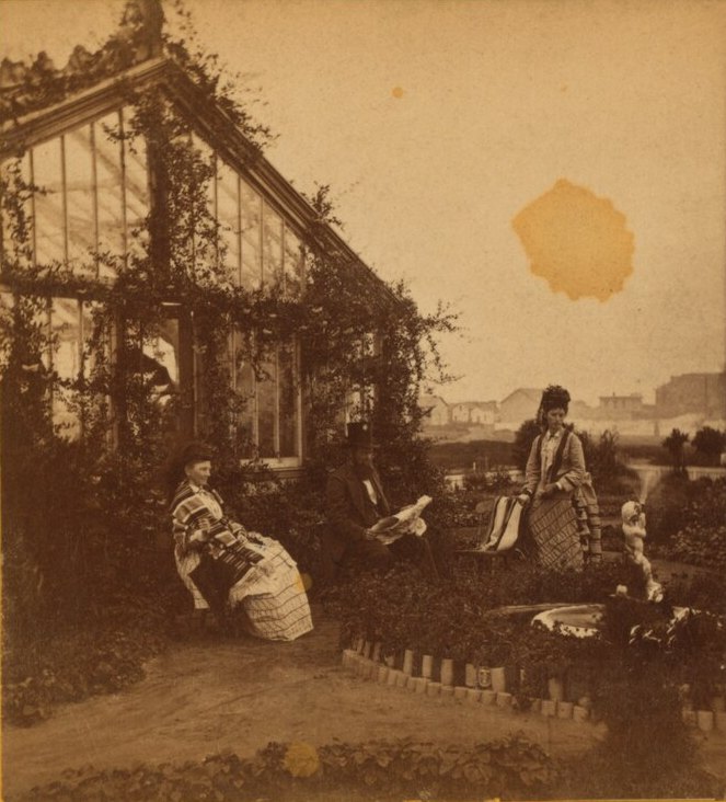 A man in a top hat reading a newspaper in a garden beside a glass greenhouse, two women nearby, low buildings on open land in the distance