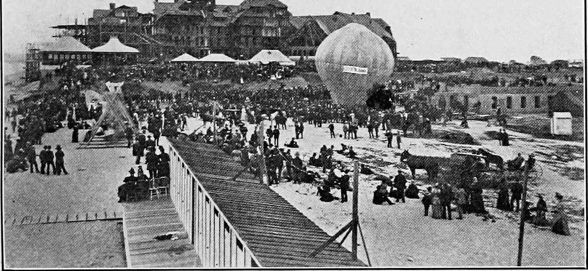 A crowd on the beach and a long boardwalk in front of the unfinished hotel, with a balloon rising over it