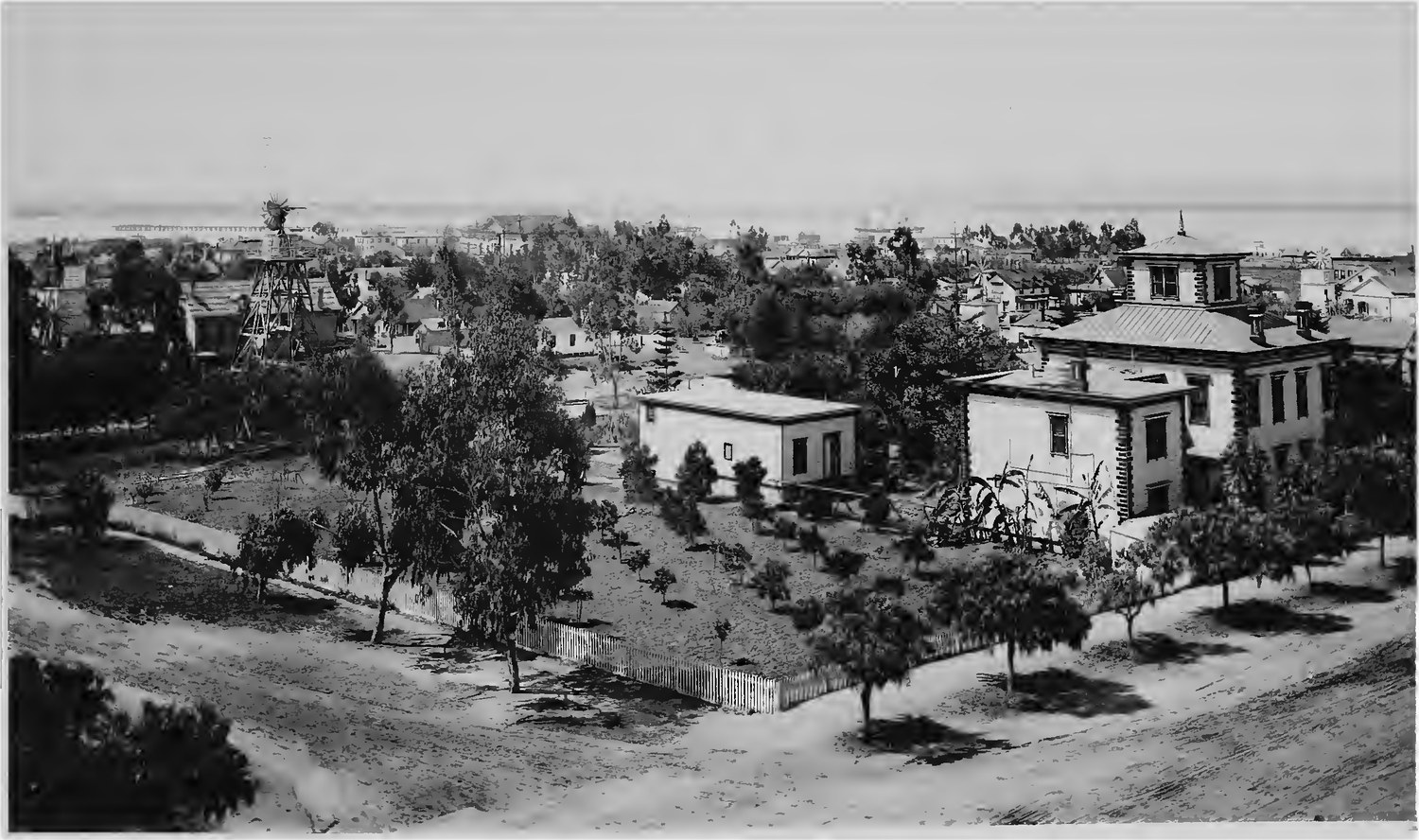 San Diego photographed in 1887 looking west toward the harbor, a windmill and water tower over scattered frame houses, young trees and a picket fence, with open dirt lots between them and Point Loma on the horizon