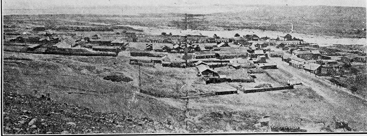 A wide view down a slope to a scattered town of low buildings, with the bay in the distance