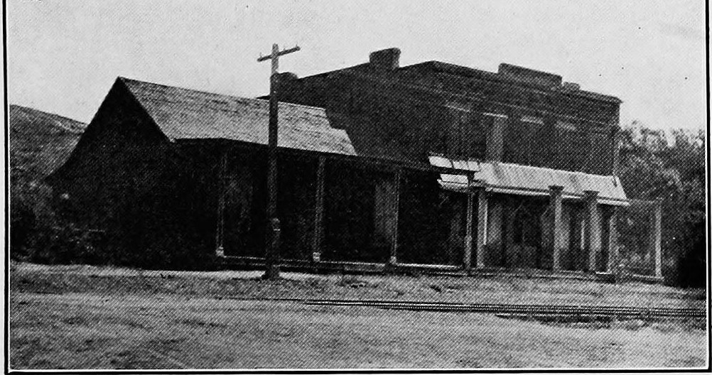 A two story brick building and a wooden storefront beside it on a dirt road, with a pole in front