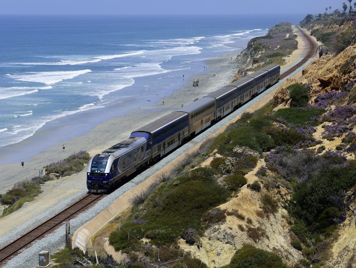 A passenger train running along tracks at the base of sandy coastal bluffs with the ocean on the left