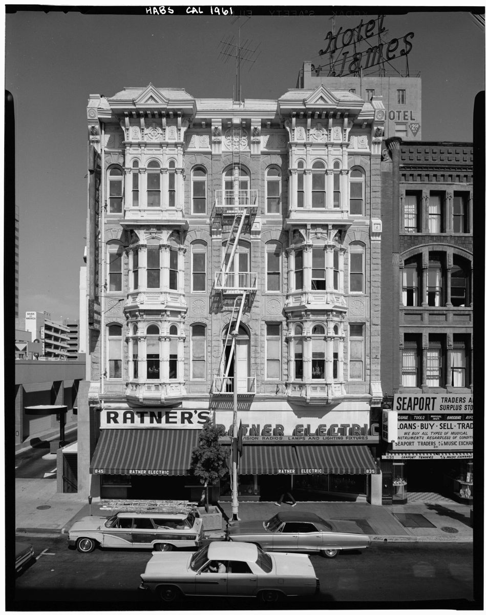 A four story Victorian building with stacked bay windows above a ground floor electric supply store, a Hotel St. James sign beside it and 1960s cars at the curb