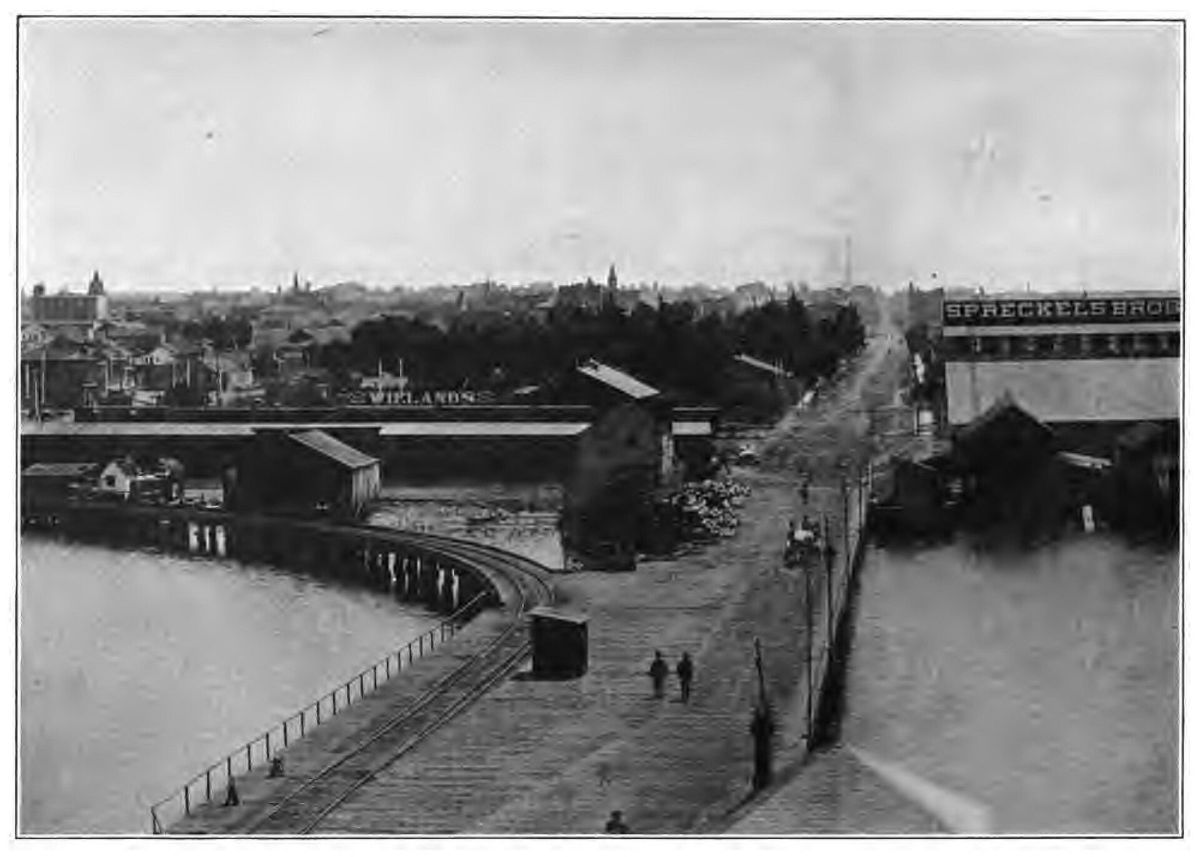 An old photograph looking down a long wooden wharf running out into the bay, warehouses on both sides and a town spreading up the hill behind