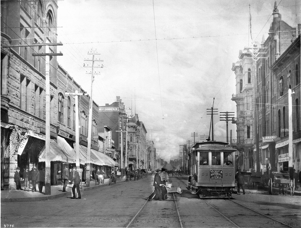 A wide downtown street with streetcar tracks down the middle, a trolley stopped at right, ornate brick and stone buildings with awnings on both sides and telegraph poles along the curb