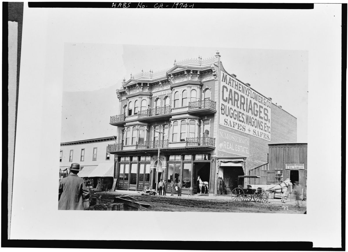 A three story Victorian hotel with stacked bay windows and iron balconies, a painted wall sign for carriages and buggies on the building beside it, a horse and wagon in the dirt street