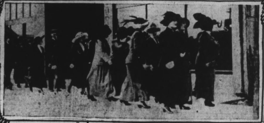 A newspaper halftone of a line of women in long coats and wide plumed hats walking along a sidewalk past a storefront, a few men among them